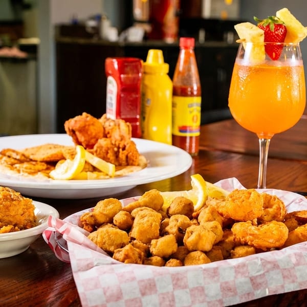 Fried seafood basket, sweet potato casserole, and a cocktail on a wooden table.