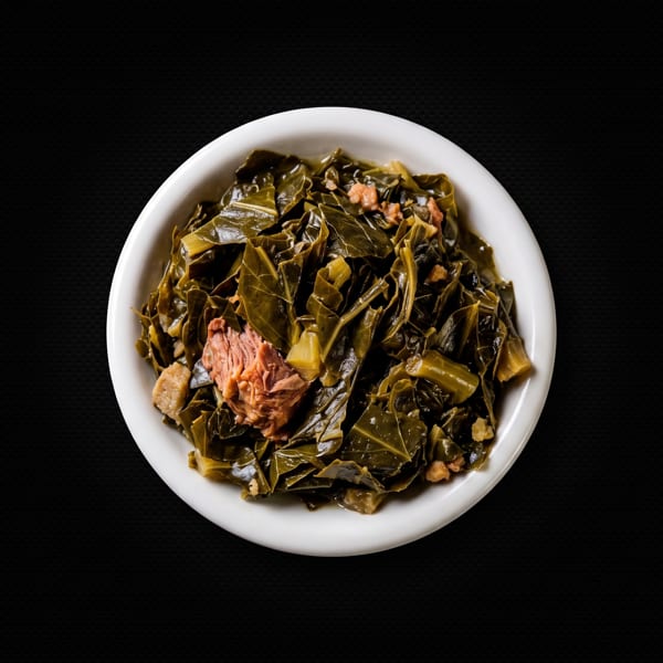 A white bowl filled with dark green collard greens and pieces of meat on a black background.