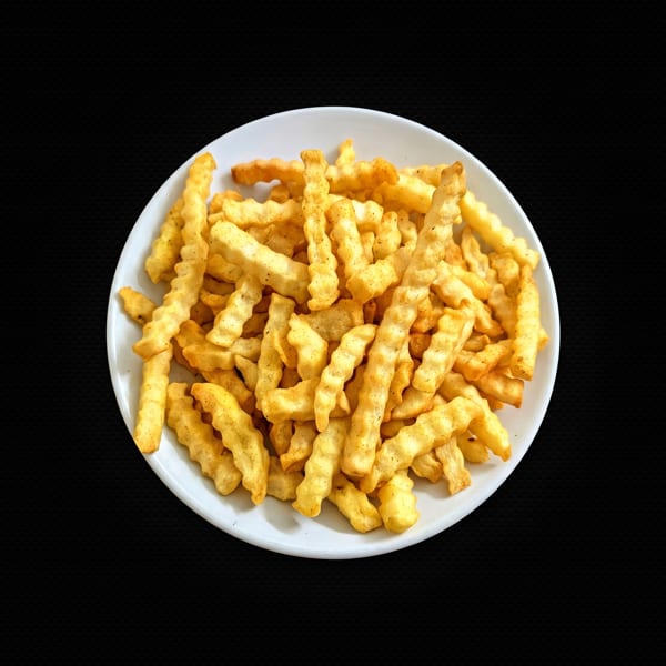 A white plate filled with golden, crispy crinkle-cut french fries, seasoned with pepper, against a black background.